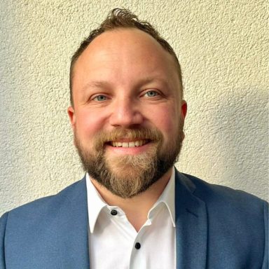 Lächelnder Mann mit kurzem Bart, blauer Jacke und weißem Hemd vor hellgrauer Wand.